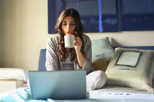 PayPal-Kosten: Frau mit Tasse in der Hand sitzt auf Bett, vor sich ein Laptop. Bild: Pexels/©Andrea Piacquadio
