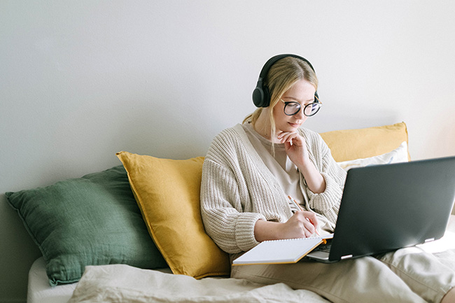 Online-Übersetzer: junge Frau auf Sofa mit Kopfhörern, Laptop und Schreibblock. Bild: Pexels/@ivan-samkov