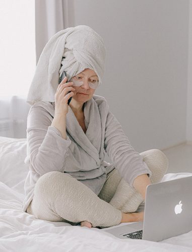 WhatsApp-Urlaubsmodus: Frau mit Handtuch auf dem KOpf und im Bademantel auf dem Bett telefoniert und hat ein Laptop vor sich. Bild: Pexels/@shvetsa