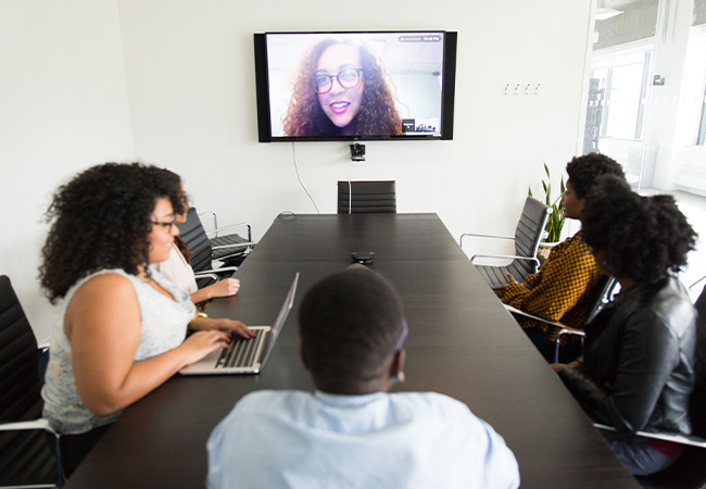 Virtuelle Konferenz: Vier Personen in Konferenzraum um einen Tisch, eine Person per Videokonferenz zugeschaltet. Bild: Unsplash/Christina @ wocintechchat.com