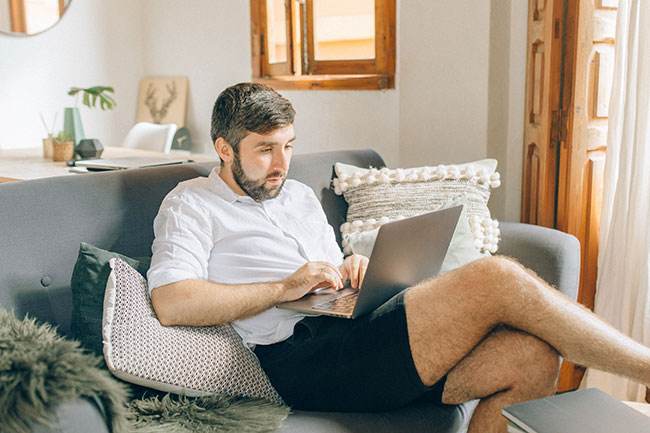 Home-Office-Pflicht: Mann in kurzer Kleidung auf dem Sofa mit Laptop. Bild: Pexels/Nataliya Vaitkevich