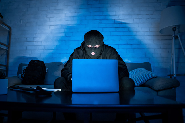 LastPass-Hack: Maskierter Hacker mit PC, der im Dunkeln auf der Couch sitzt und große Daten stiehlt. Bild: ©Prostock-studio/stock.adobe.com