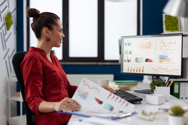 Lächelnde Geschäftsfrau in rotem Oberteil sitzt am Schreibtisch im Büro. Bild: stock.adobe.com/DC Studio (https://stock.adobe.com/de/images/businesswoman-with-clipboard-sitting-at-desk-in-startup-business-office-smiling-employee-in-red-shirt-comparing-charts-successful-entrepreneur-looking-at-desktop-computer-to-analyze-business-data/473777212?asset_id=473777212)