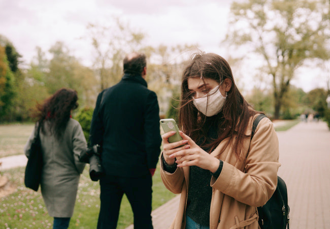 Eine Frau schaut verträumt auf ihr Handy während hinter ihr ein Paar spazieren geht. Fällt Sie auf Social Hacking rein? Bild: Pexels/Maksim Goncharenok