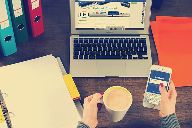 Eine Person hält ihr Smartphone in der rechten und eine Tasse Kaffee in der linken Hand. Sie sitzt an einem Schreibtisch mit verschiedenen Ordnern und einem aufgeklappten Laptop. Gerade in den sozialen Netzwerken, die auf den beiden Bildschirmen zu sehen sind, sollten Sie auf Netiquette und das Urheberrecht achten. Pixabay/William Iven